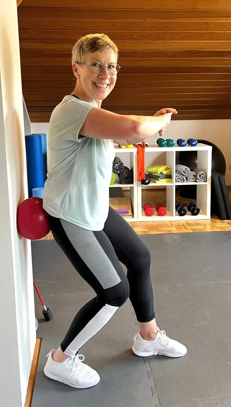Suse Wirtherle in ihrem Privat-Studio in Bammental – sie lächelt in die Kamera, im Hintergrund die Holzdecke und das Regal mit Faszienrollen, Bändern und Hanteln